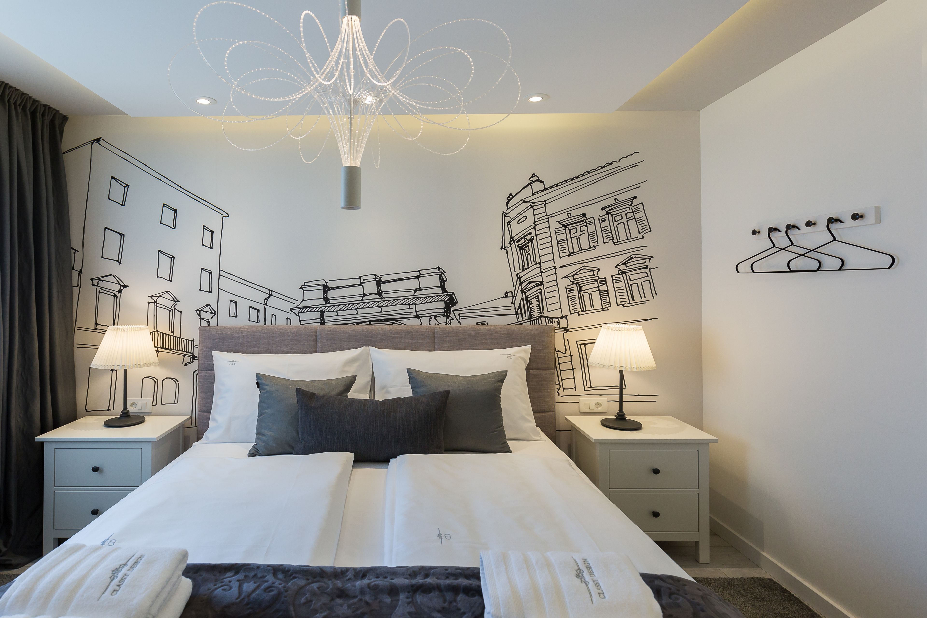 Habitación de hotel con una cama grande, dos mesitas de noche con lámparas, un candelabro y arte mural que representa edificios.