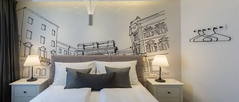 Habitació d'hotel amb un llit gran, dues taules de nit amb làmpades, un penjoll i obra d'art mural que representa edificis.