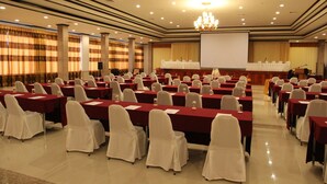 Un salón de conferencias con filas de mesas y sillas, una pantalla de proyector y una zona de escenario.