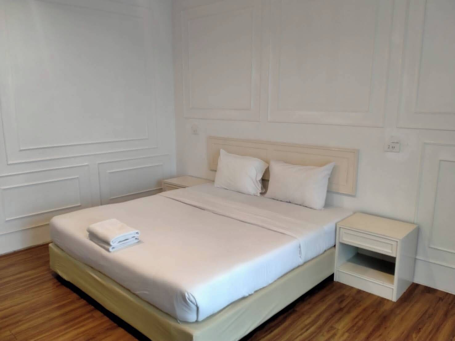 Una cama bien hecha con edredón blanco y almohadas, un cabecero de madera y una mesita de noche con cajón.
