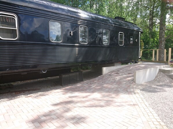 A stationary train car with multiple windows and a metal door, parked on a paved area with trees in the background. - Tågvagnshotellet - Silleruds Station (Årjäng)