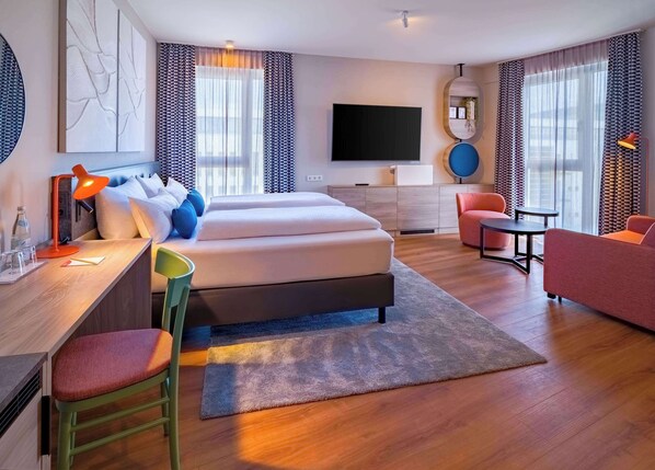 Una habitación de hotel moderna con una cama grande, un escritorio con una silla, un televisor y un área de descanso con una mesa y sillas.