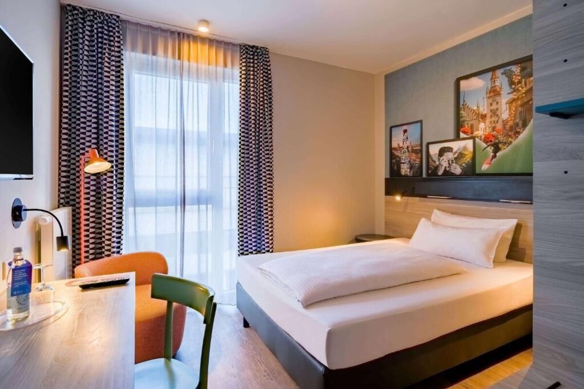 Habitación de hotel con cama, escritorio, silla, televisor y ventana con cortinas.