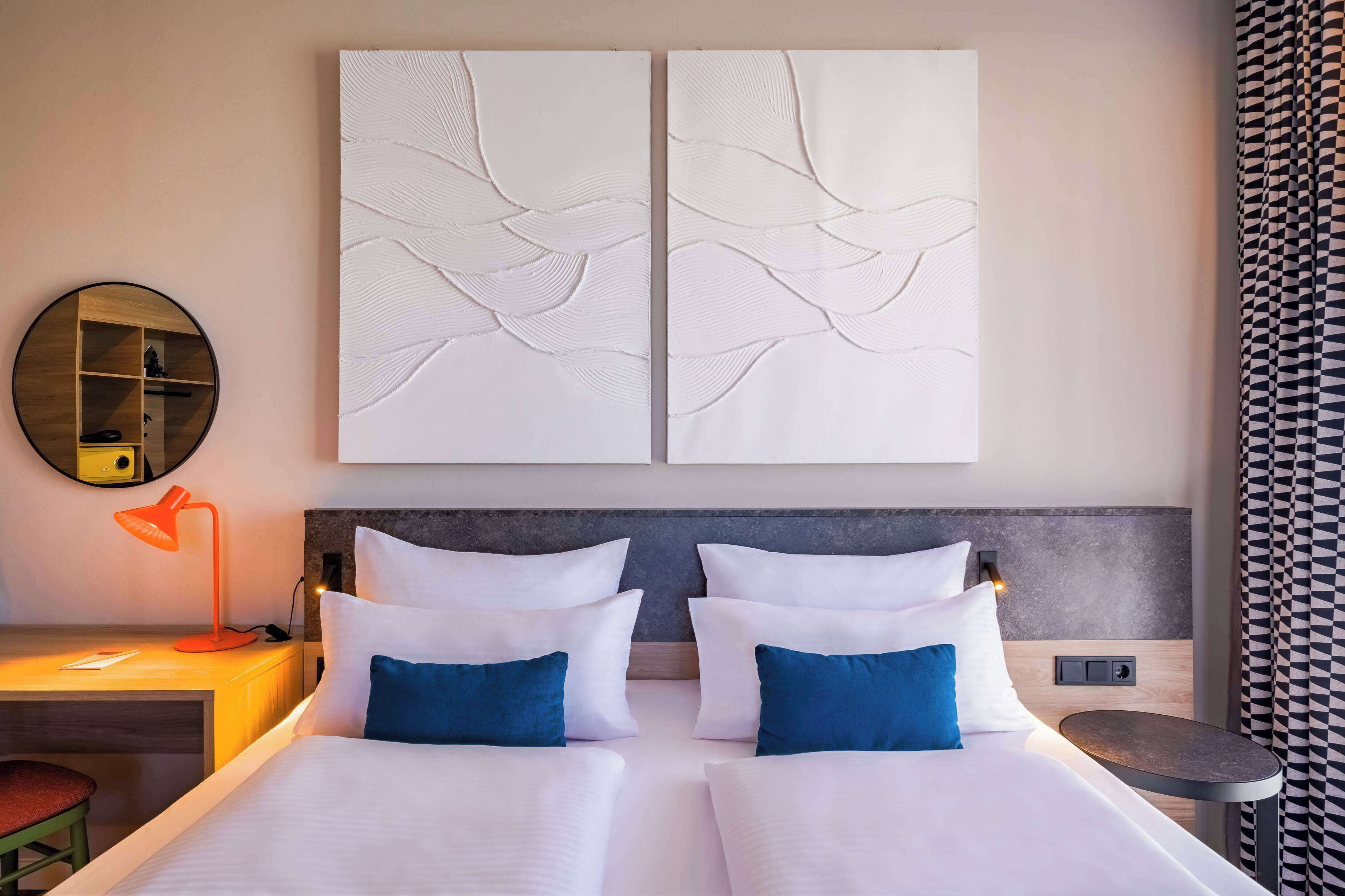 Una habitación de hotel moderna con una cama doble, dos mesitas de noche, un escritorio con lámpara y un espejo redondo.