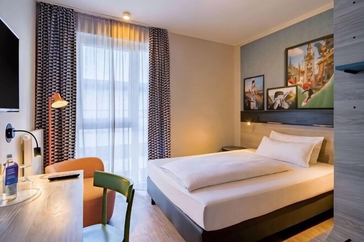 Habitación de hotel con cama, escritorio, silla, televisor y cortinas.