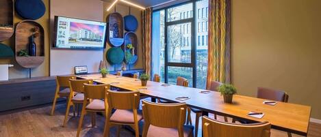 Una sala de reuniones moderna con una larga mesa de madera, sillas cómodas, un amplio ventanal con vistas a edificios y una pantalla montada en la pared que muestra una imagen de un edificio.