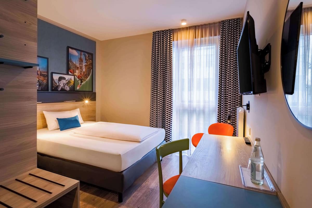 A hotel room with a bed, a desk with a chair, a TV, and a window with curtains.