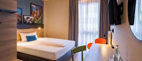 Habitación de hotel con una cama, un escritorio con silla, un televisor y una ventana con cortinas.