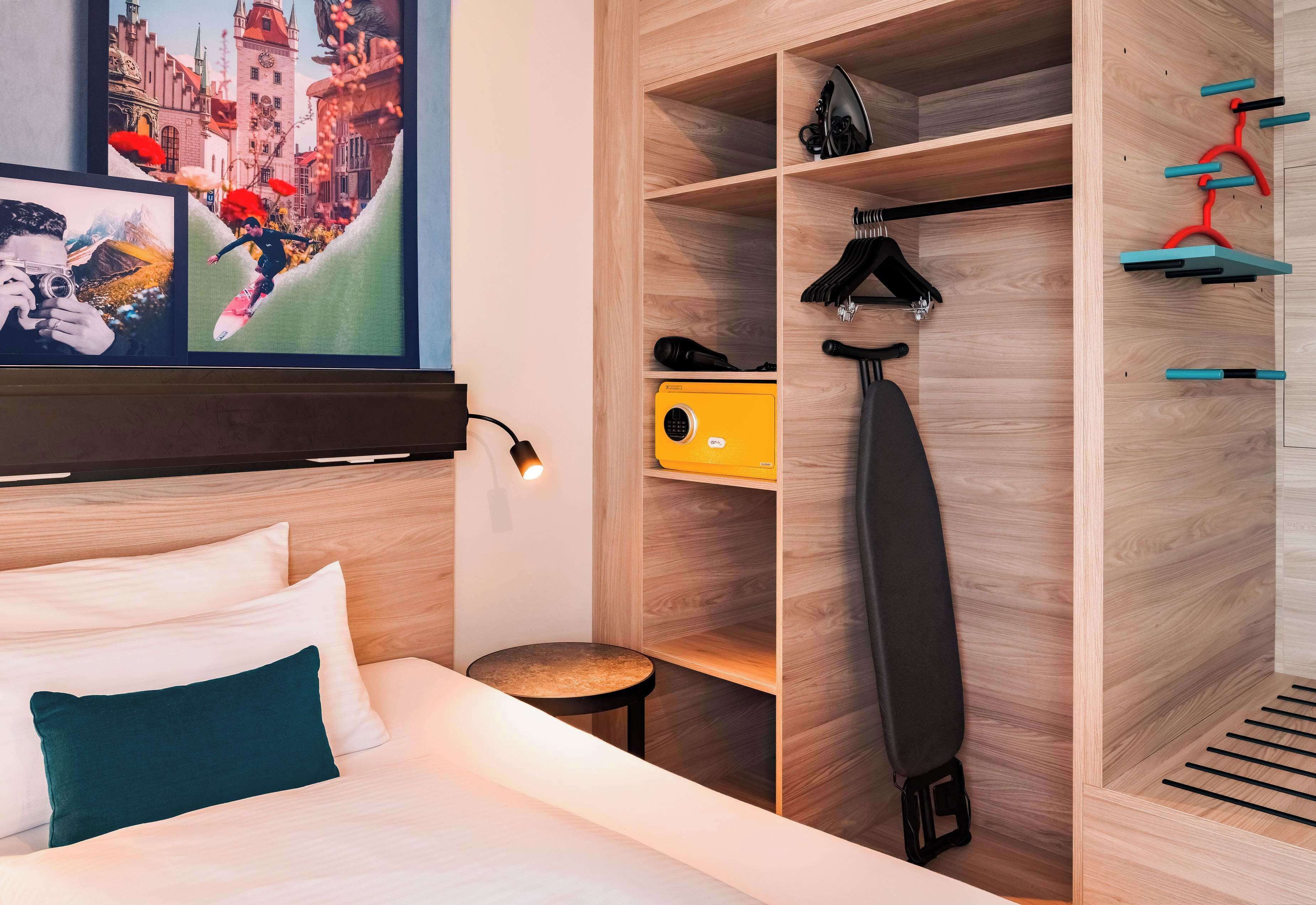 A hotel room with a bed, a round table, a wardrobe with hanging space, and a yellow suitcase.