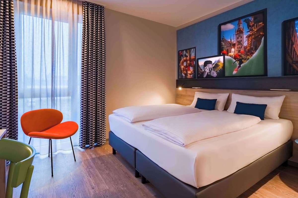 A hotel room with a large bed, a desk, and a chair. There are two paintings on the wall.