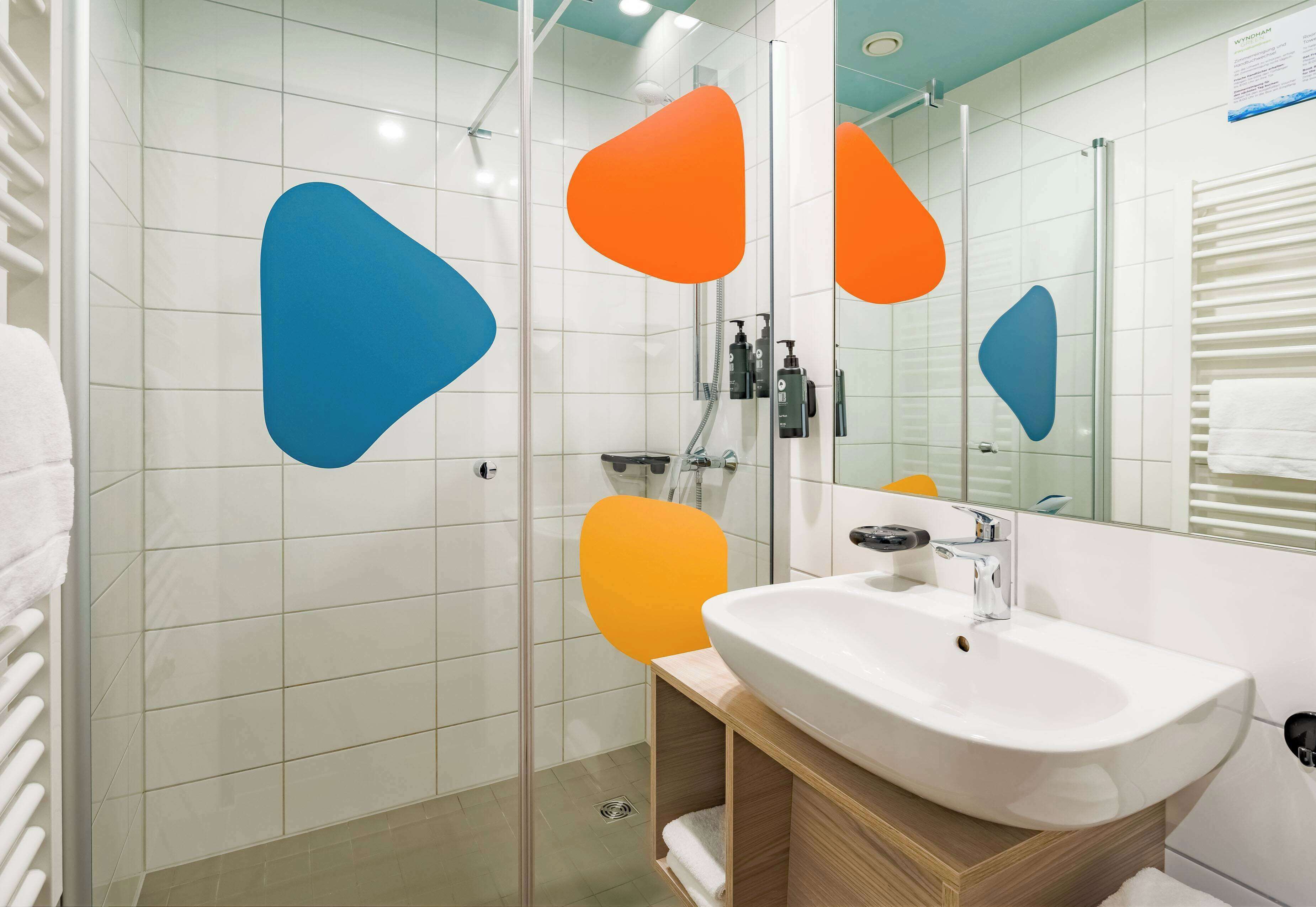 A modern bathroom with a glass shower enclosure, a white sink, and a mirror with orange and blue geometric decorations.