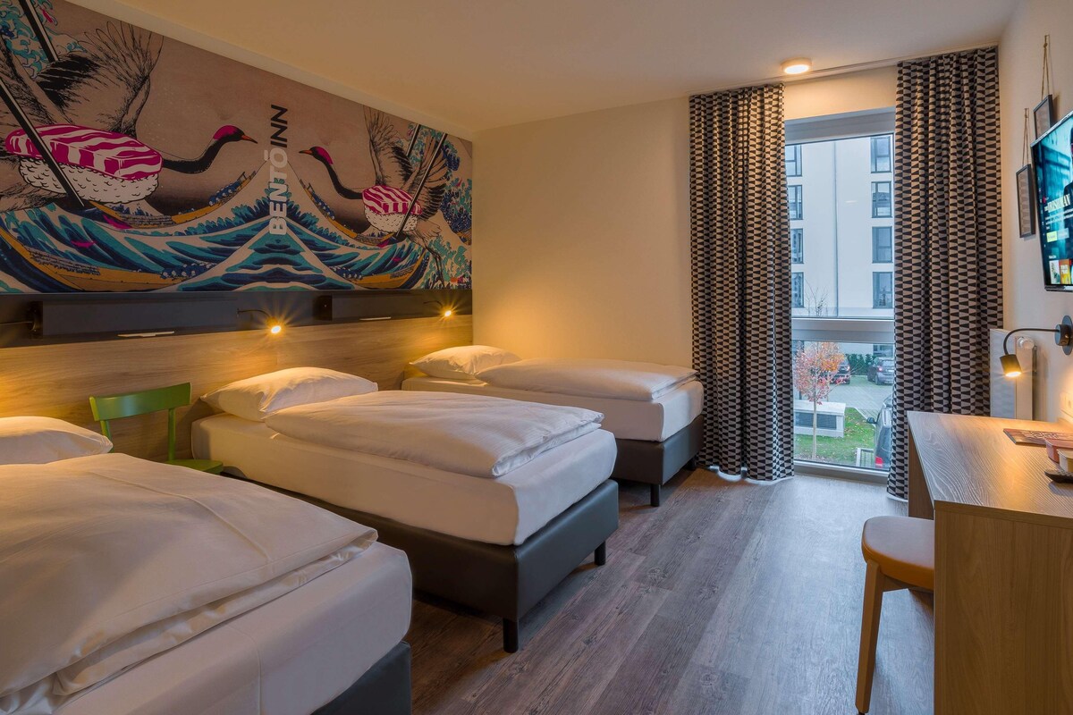 Habitación de hotel con tres camas, un escritorio y un ventanal grande con cortinas.
