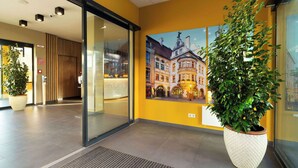 Un moderno vestíbulo de hotel con una planta grande en maceta, una entrada por puerta de cristal y un mural de un edificio histórico.