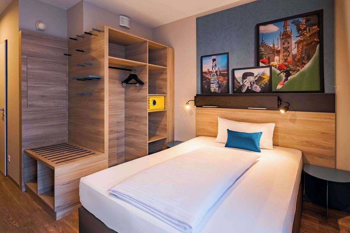 A hotel room with a bed, a wooden wardrobe, and a wall-mounted TV displaying cityscape images.