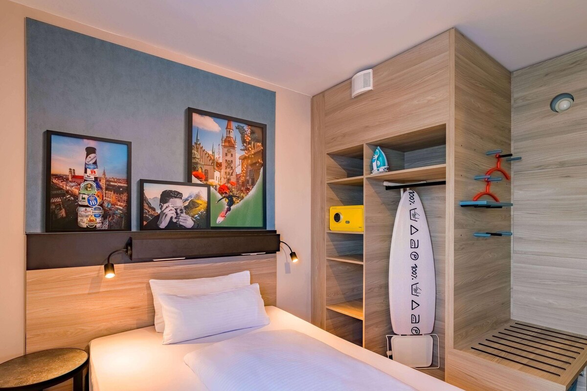 Habitación de hotel con cama, una mesa redonda y obras de arte en la pared que muestran paisajes urbanos y una escena de montaña.