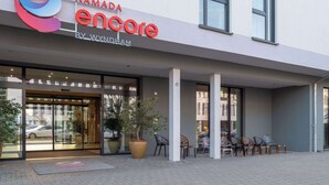 Entrada de hotel con diseño moderno, que incluye un logotipo, una placa con el nombre y un área de estar con sillas afuera.