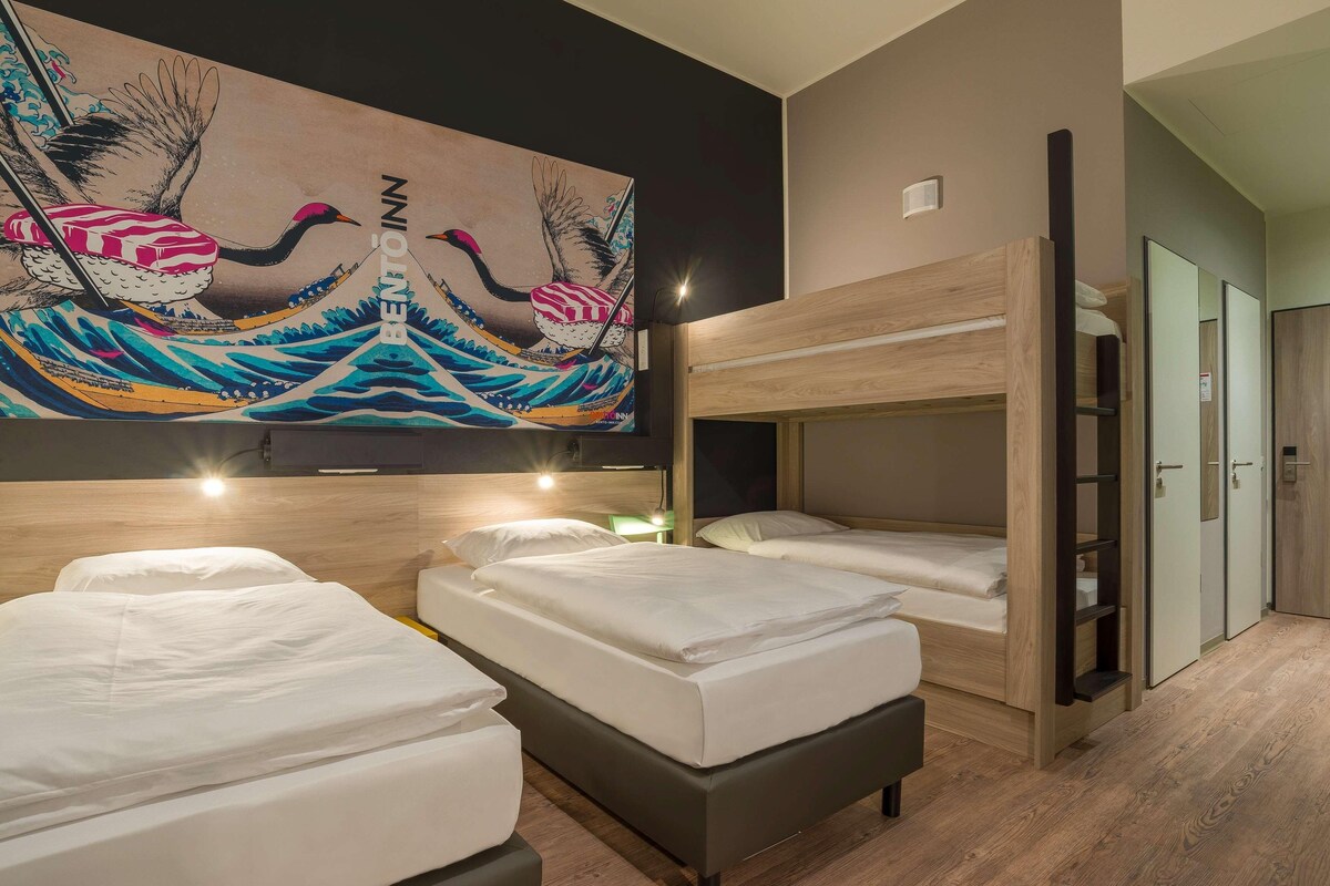 Habitación de hotel con dos camas, una litera, un cuadro grande en la pared y piso de madera.