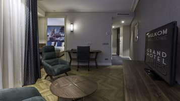 A modern hotel room with a wooden herringbone floor, a round wooden table, two green armchairs, a dark armchair, a flat-screen TV, and a 'Welcome Grand Hotel' sign.