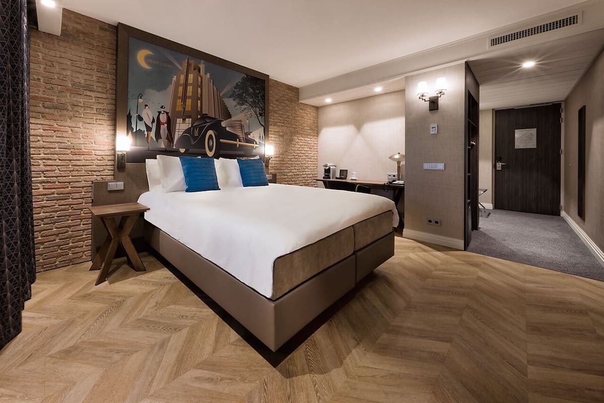 Ein modernes Hotelzimmer mit einem großen Bett, einer Wandmalerei, einem Schreibtisch mit Stuhl und einer Tür zu einem weiteren Raum.