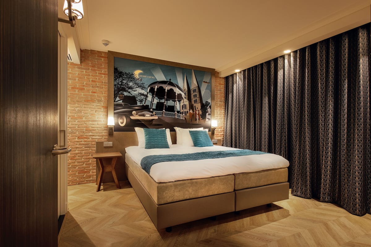 Ein modernes Hotelzimmer mit einem großen Bett, einem Holz-Nachttisch, einer Ziegelwand und einem Wandgemälde einer Stadtansicht.