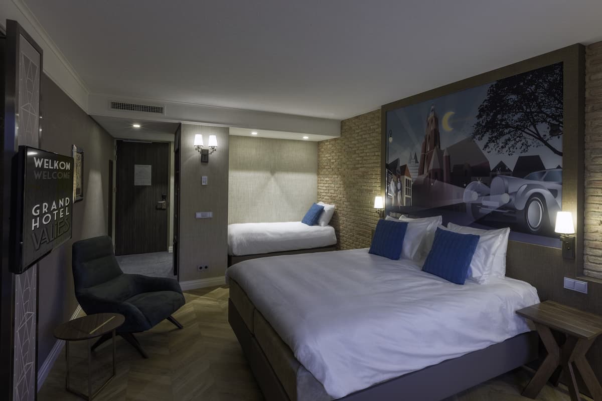 Ein Hotelzimmer mit einem großen Bett, einem kleineren Bett, einem Sessel und einem Schreibtisch. An der Wand hängt ein Gemälde und ein Schild mit der Aufschrift 'Welcome Grand Hotel'.