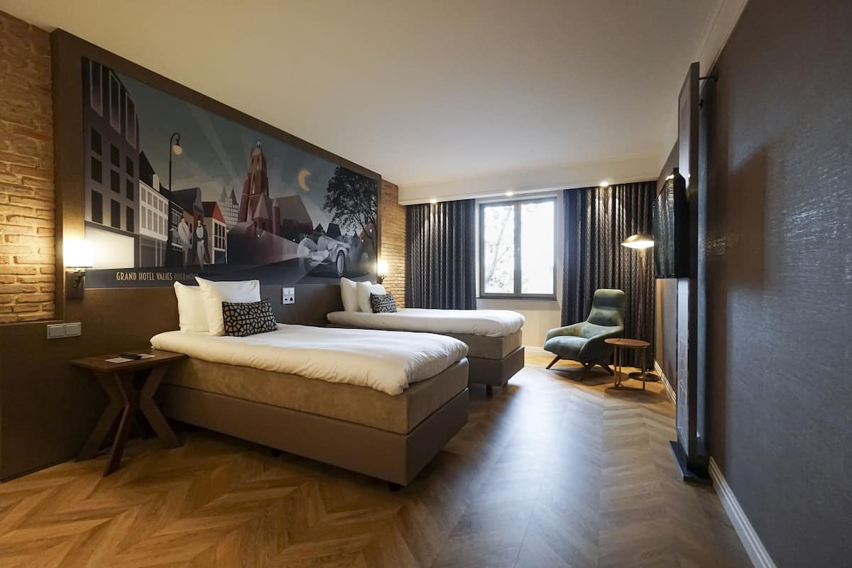 Ein Hotelzimmer mit einem großen Bett, einem kleineren Bett, einem Sessel, einem Tisch und einem Wandgemälde.