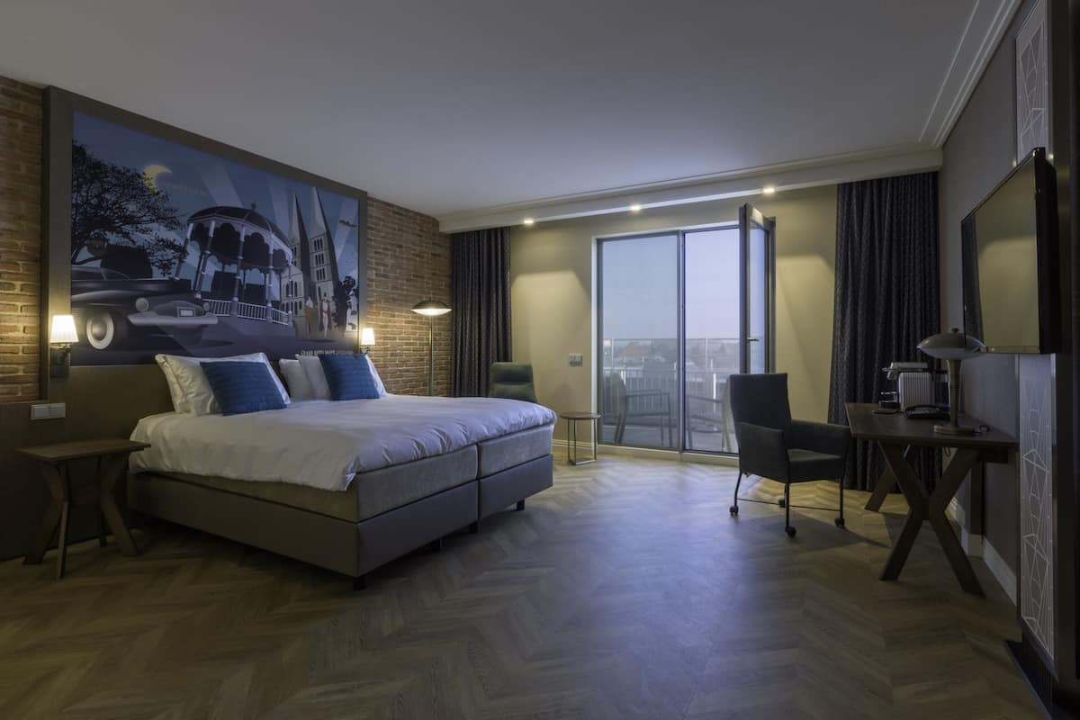 Ein modernes Hotelzimmer mit einem großen Bett, einem Schreibtisch, einem Sessel und Blick auf die Stadt.