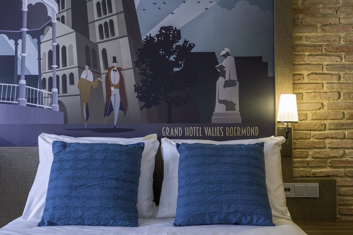 Ein Hotelzimmer mit einem Bett, zwei blauen Kissen, einer weißen Tagesdecke, Wandgemälden und einer Ziegelwand.