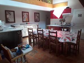 A kitchen with a dining area, featuring a tiled floor, wooden chairs, a table with a patterned tablecloth, and a refrigerator.
