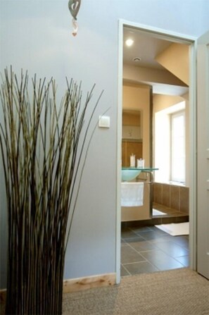 A hallway with a tall plant, a glass bowl on a shelf, and a bathroom visible through an open door.