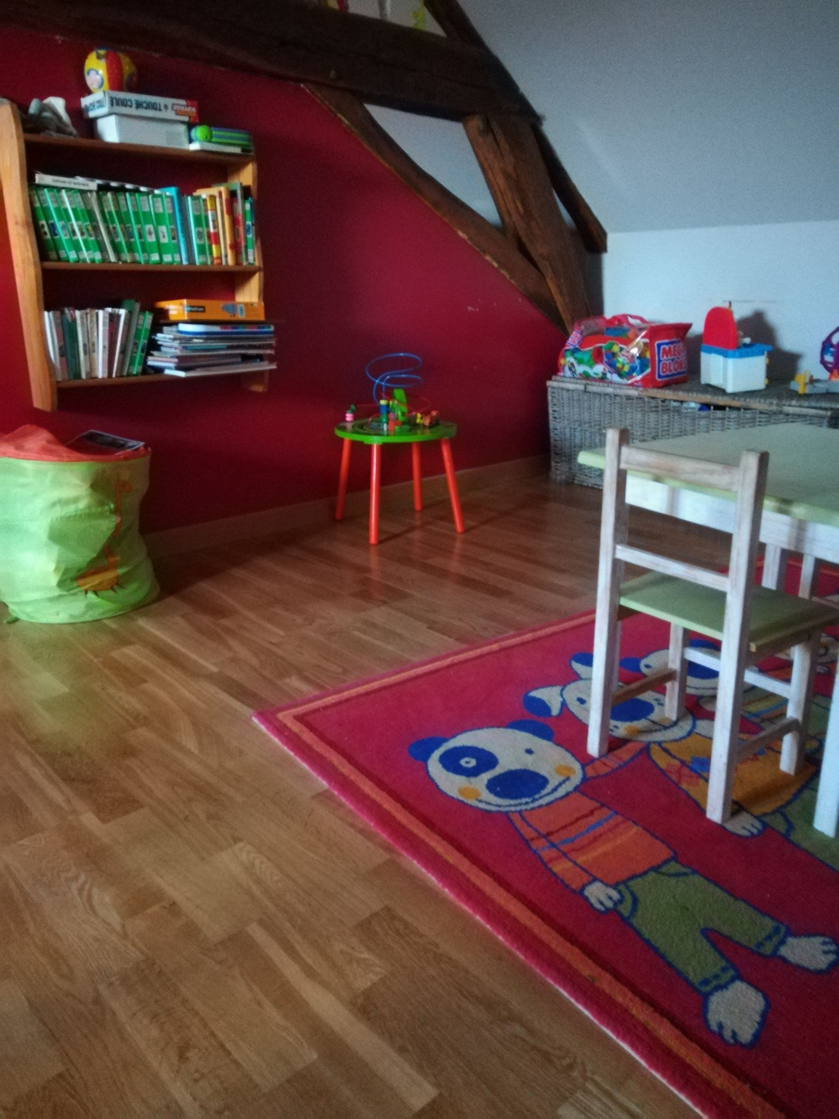 A room with a wooden floor, a red wall, a bookshelf, a small green table, a red rug with a cartoon character, and a white chair.