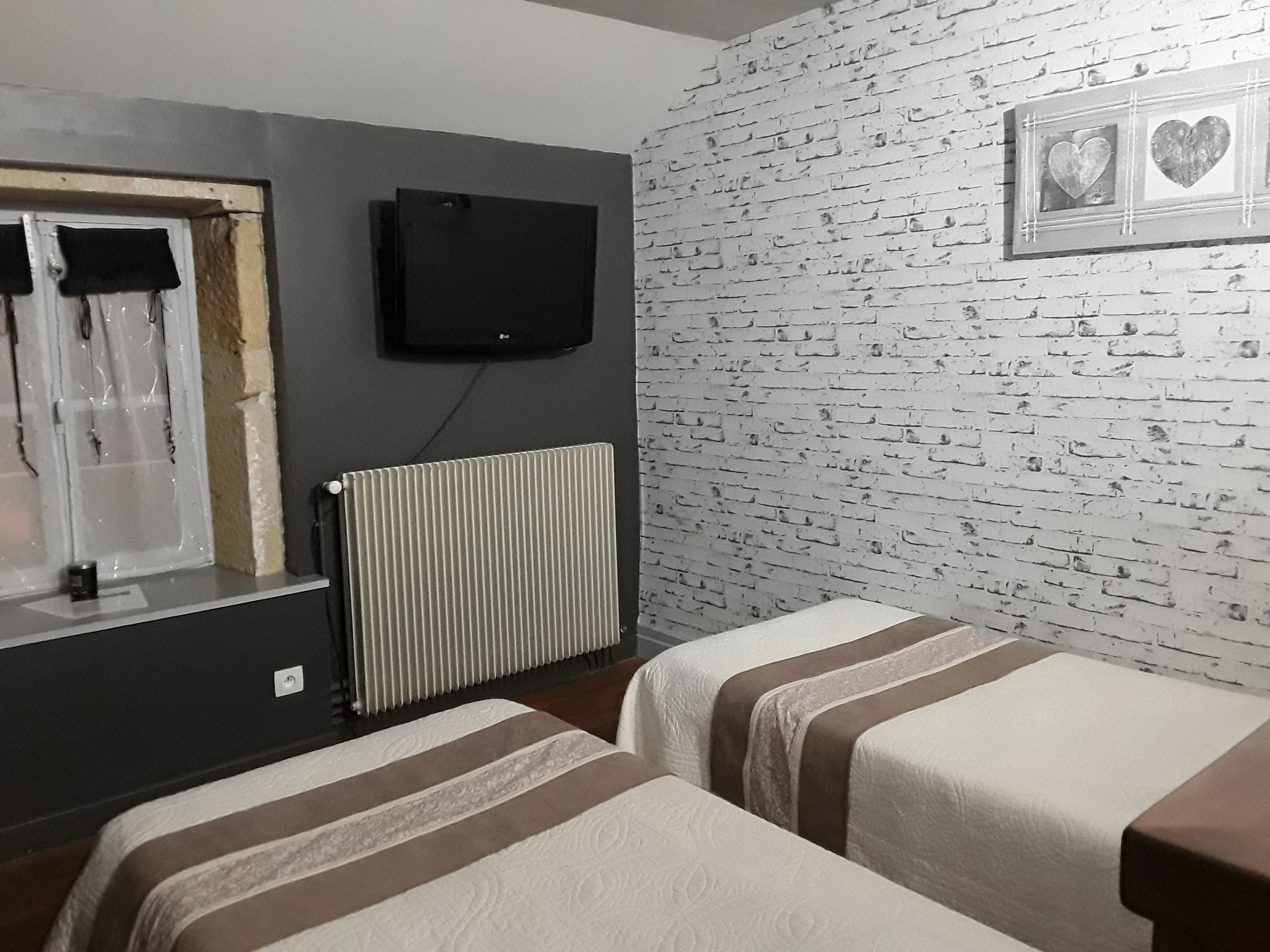 Una habitación con dos camas, un radiador, un televisor montado en la pared y una ventana con cortinas.