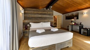 A modern bedroom with a wooden ceiling, a large bed with white linens, a nightstand, a desk with a lamp, and a painting on the wall. - Du Parc Hotel (Sauze d'Oulx)