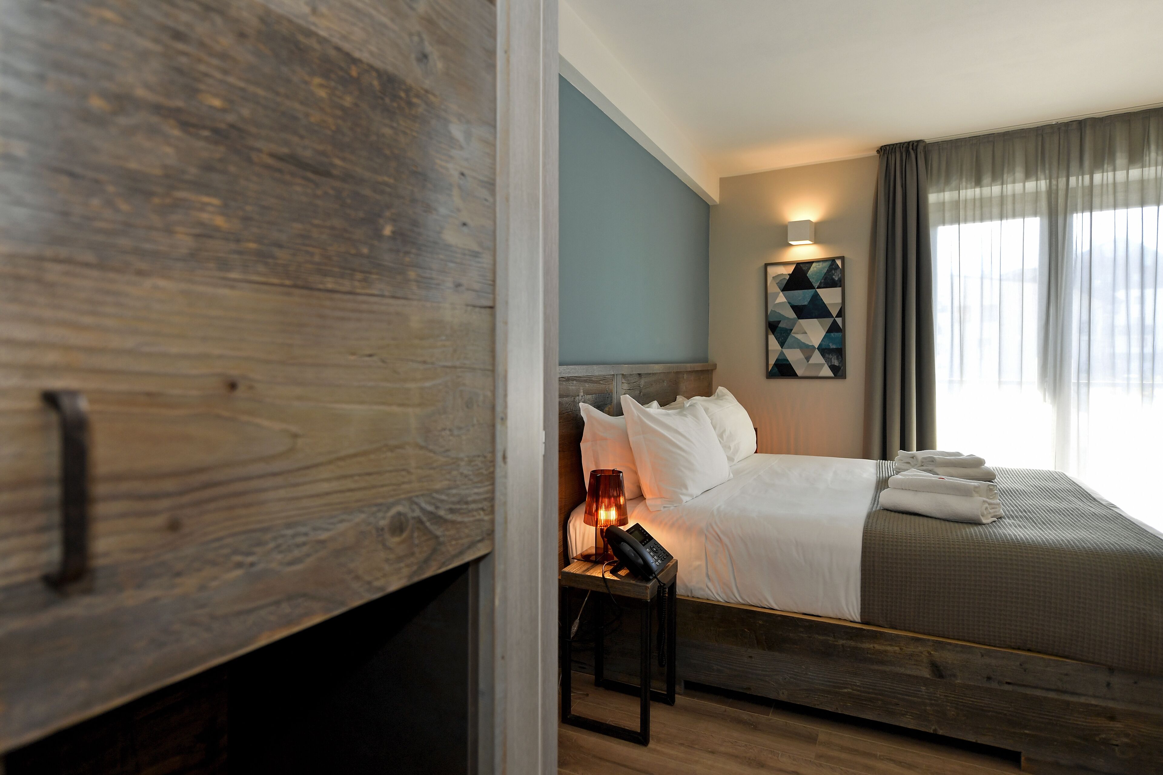 A modern hotel room with a wooden headboard, a bedside table with a lamp, and a bed with neatly folded towels.
