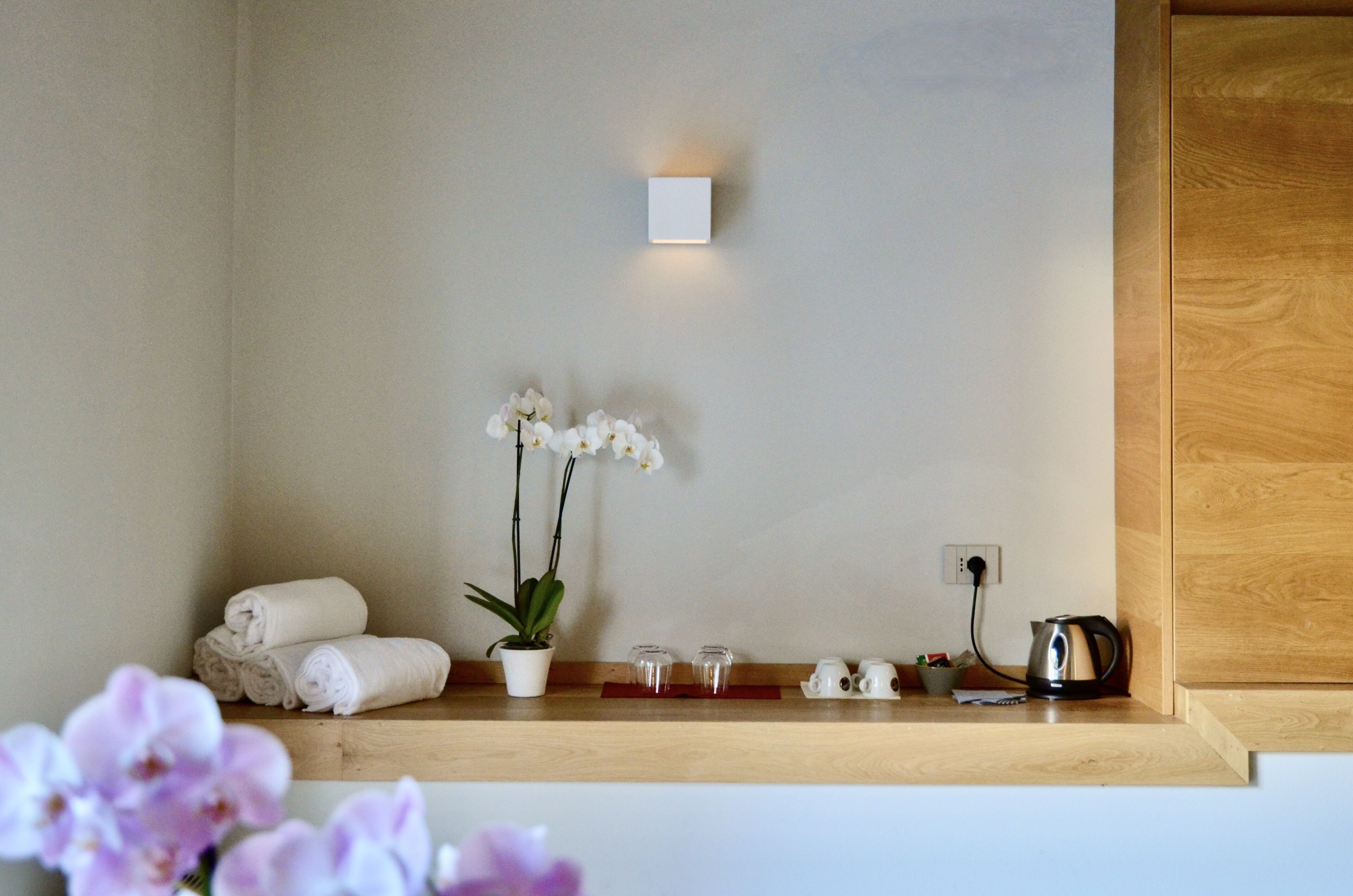 Une salle de bain avec une étagère en bois où sont posés des serviettes, une orchidée en pot et une bouilloire.