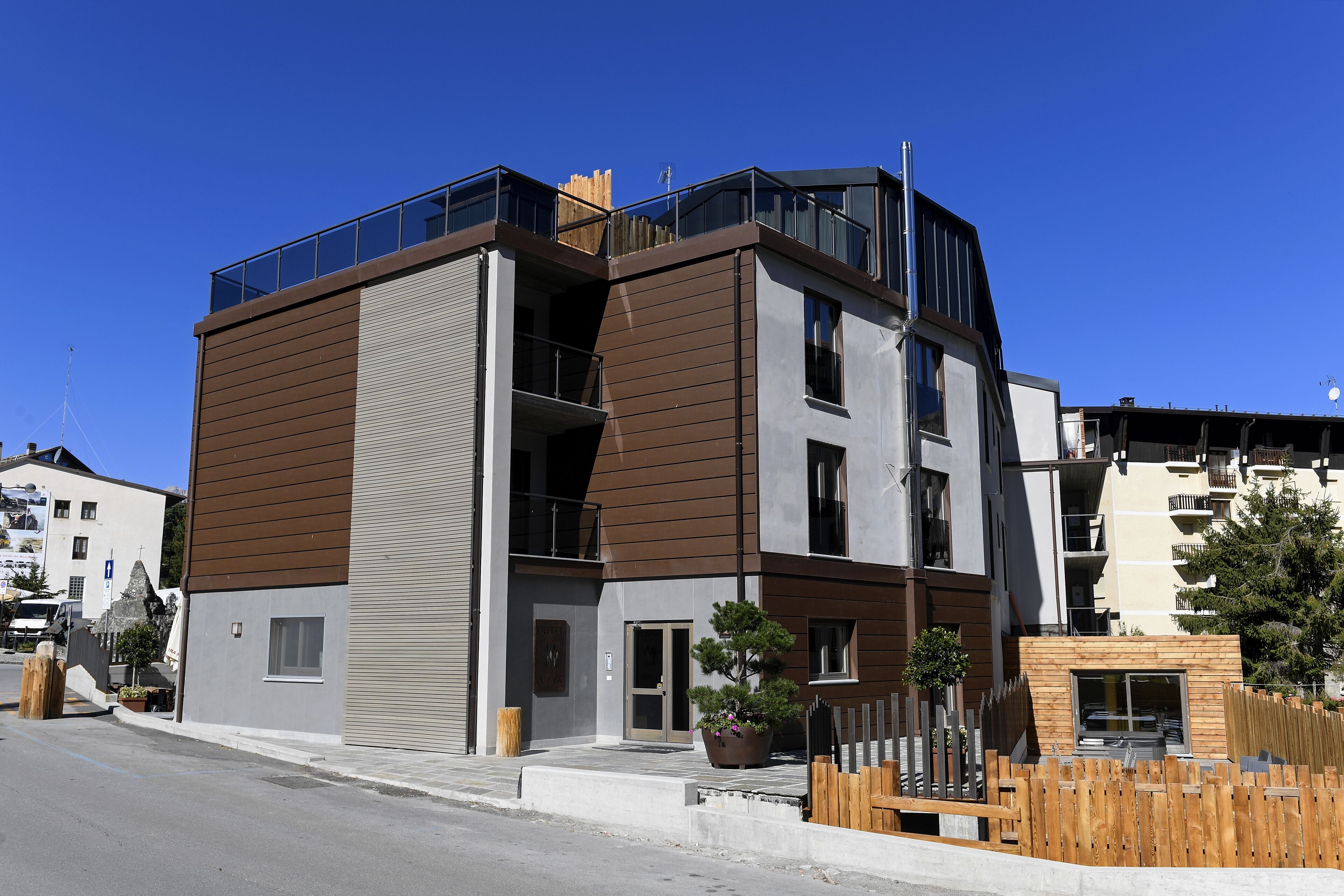 A modern multi-story building with a mix of brown and grey exterior panels, large windows, and a balcony on the upper floor.