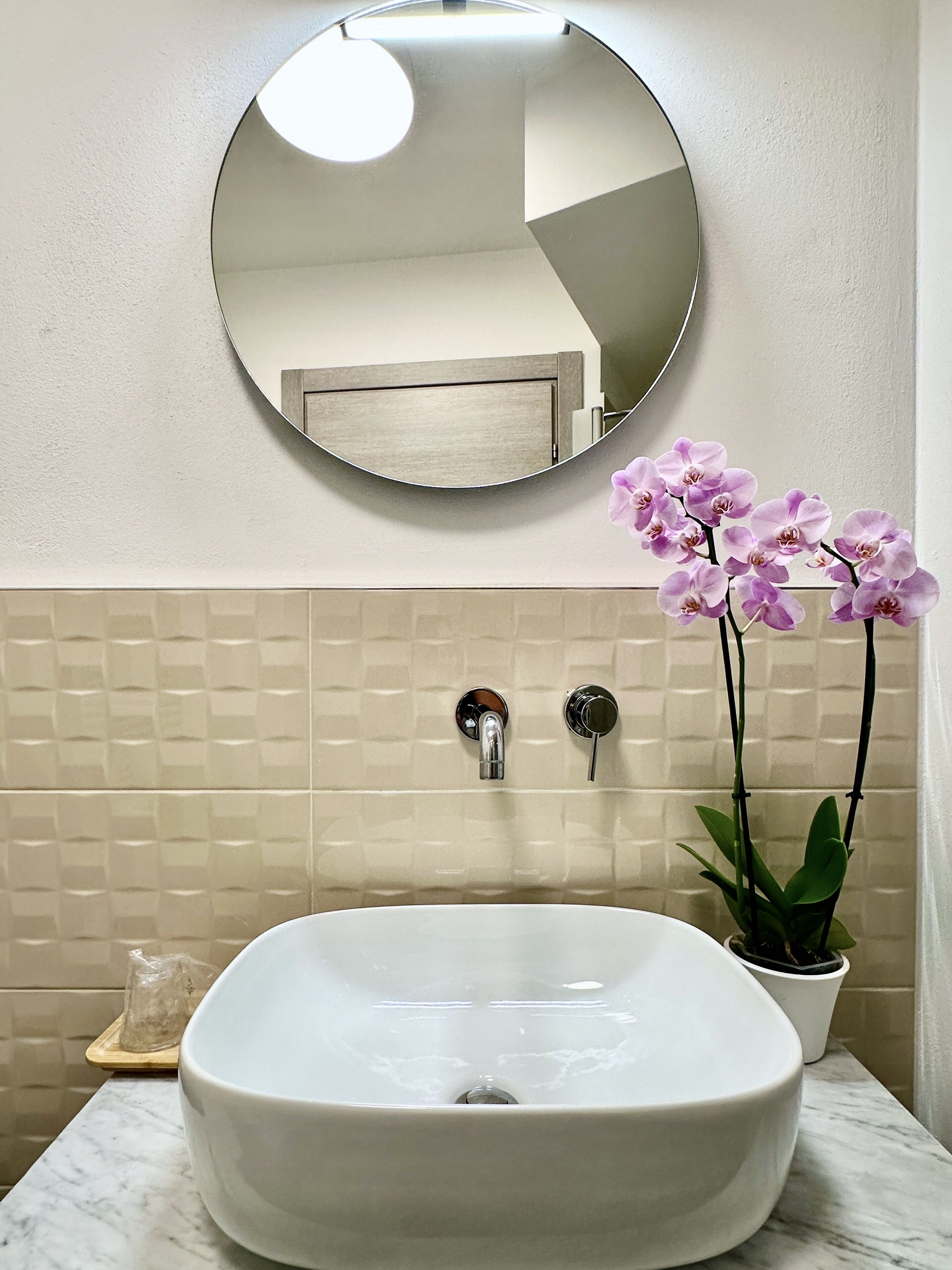 Un baño con un espejo redondo, un lavabo blanco y una orquídea en maceta.