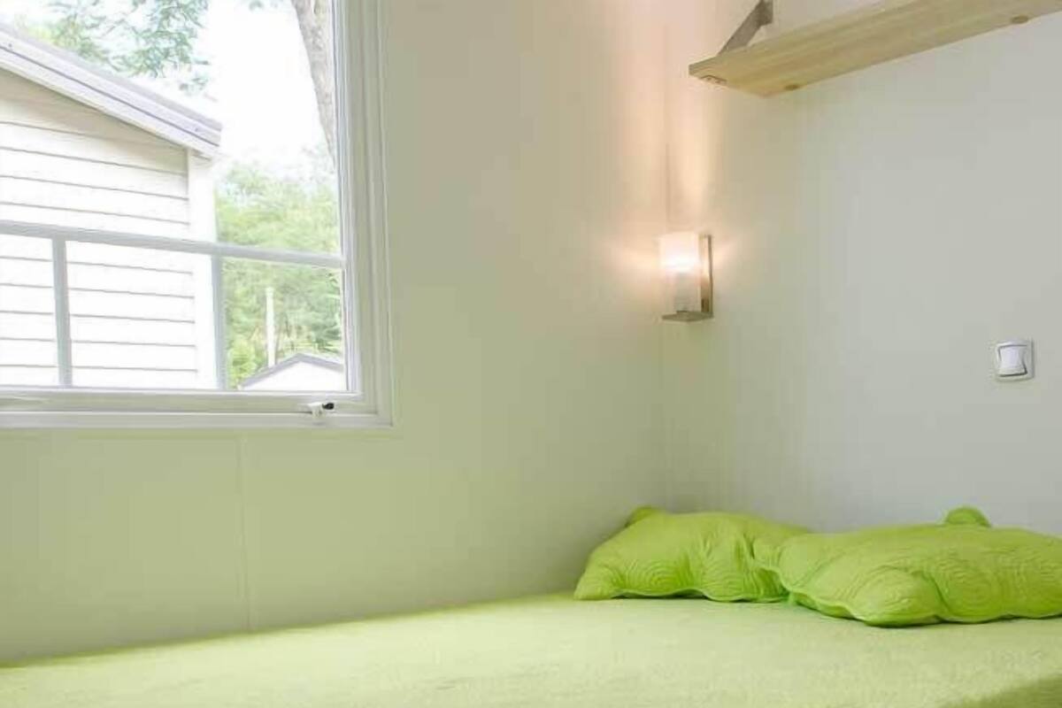 A small, single-bed room with a window, a wall-mounted light fixture, and a green bedspread.