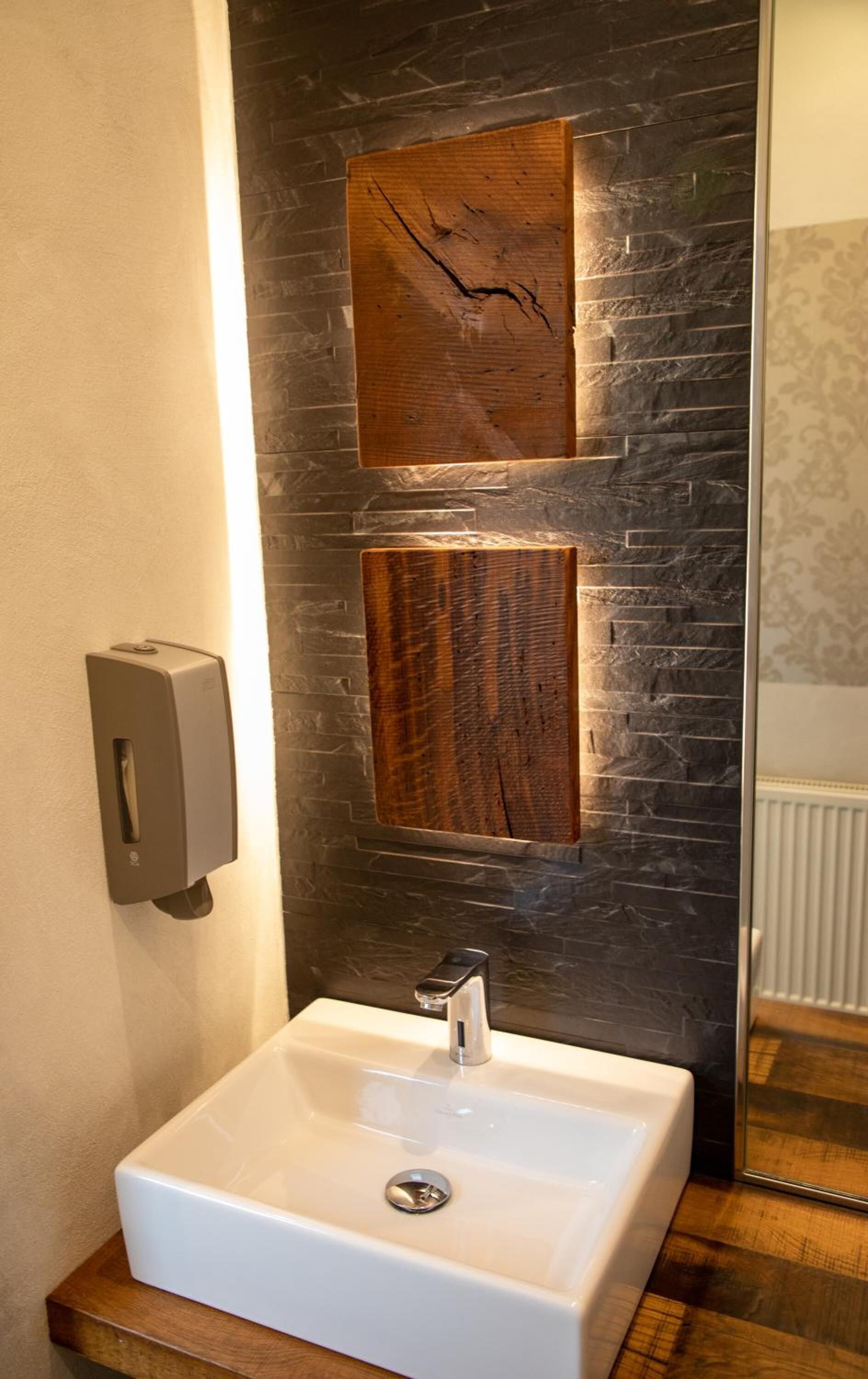 Ein modernes Badezimmer mit einem weißen Waschbecken, zwei Holzwänden und einem Handdesinfektionsspender.