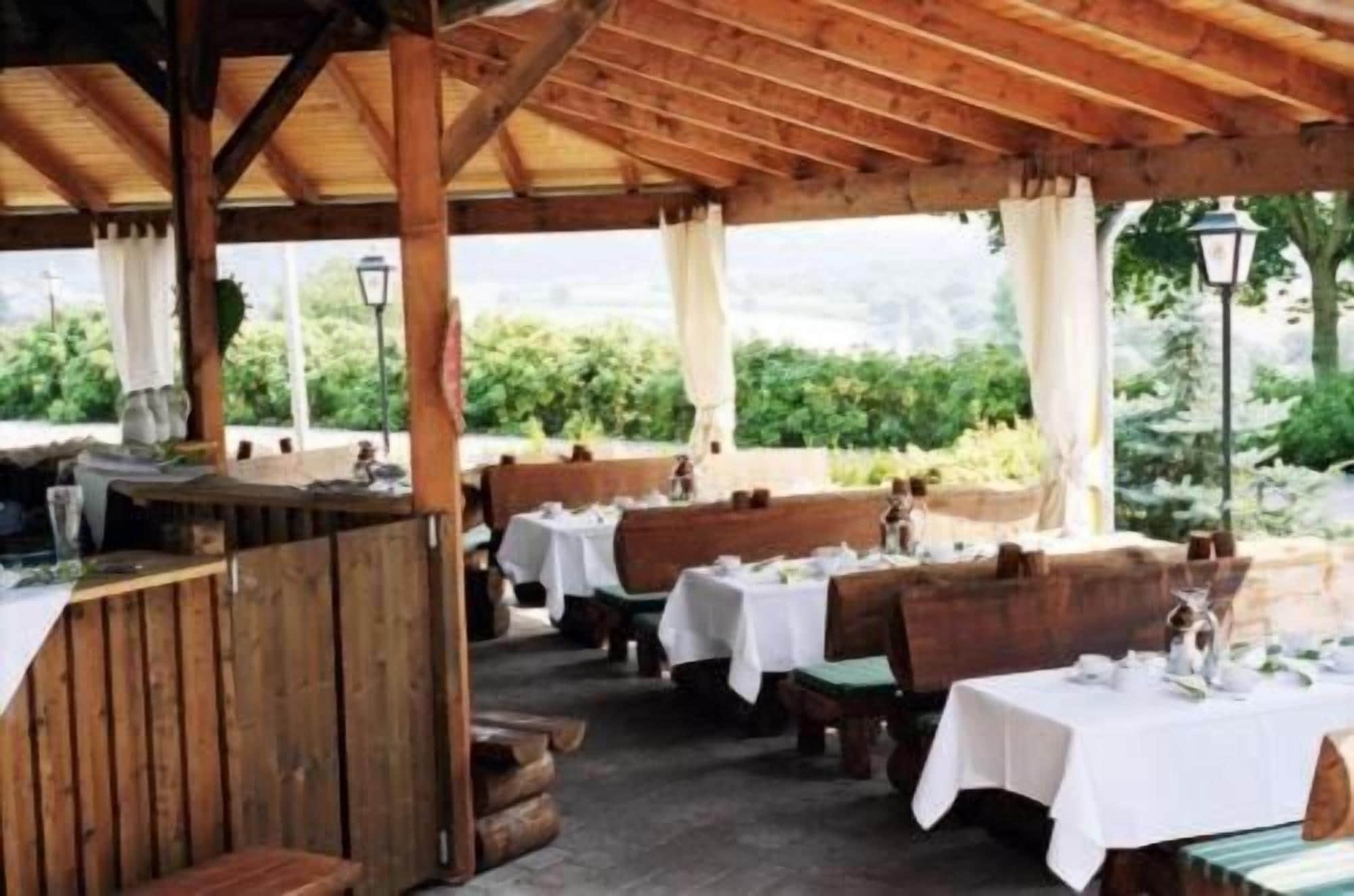 Un restaurante al aire libre con mesas y bancos de madera, cubierto por un techo de madera, con un fondo de vegetación.