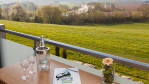 A balcony with a clear view of a green field and trees. There is a bottle, two glasses, a pair of glasses, a pen, and a small vase with a flower on a table. - Landhotel Weserblick (Beverungen)