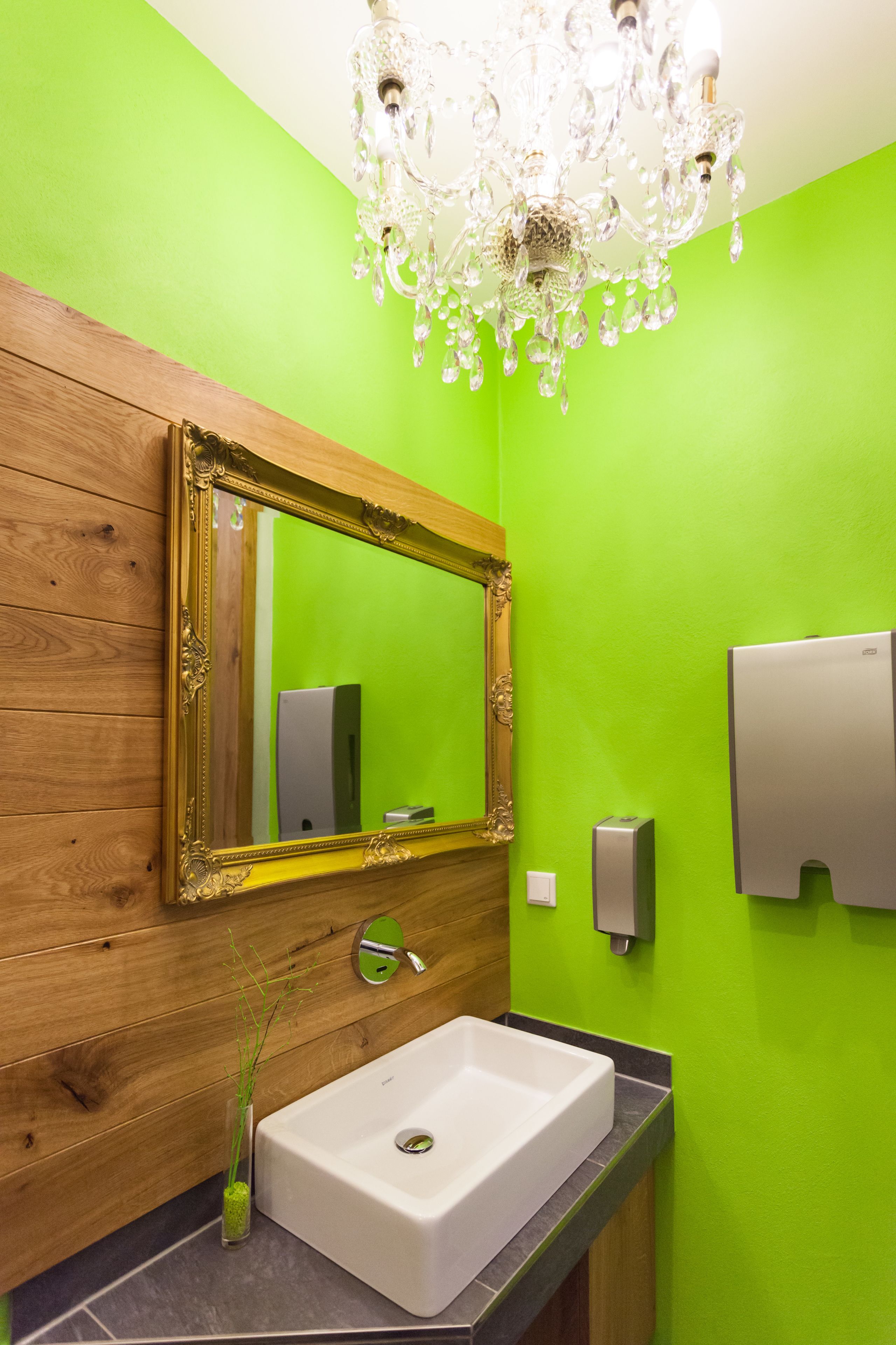 A bathroom with a wooden vanity, a white sink, a mirror with an ornate frame, a chandelier, and a green wall.