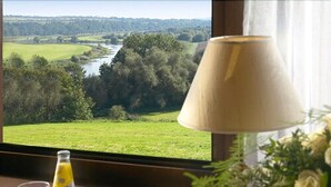 A room with a view of a river and greenery, a table with glasses and a bottle, and a lamp with a flower arrangement. - Landhotel Weserblick (Beverungen)