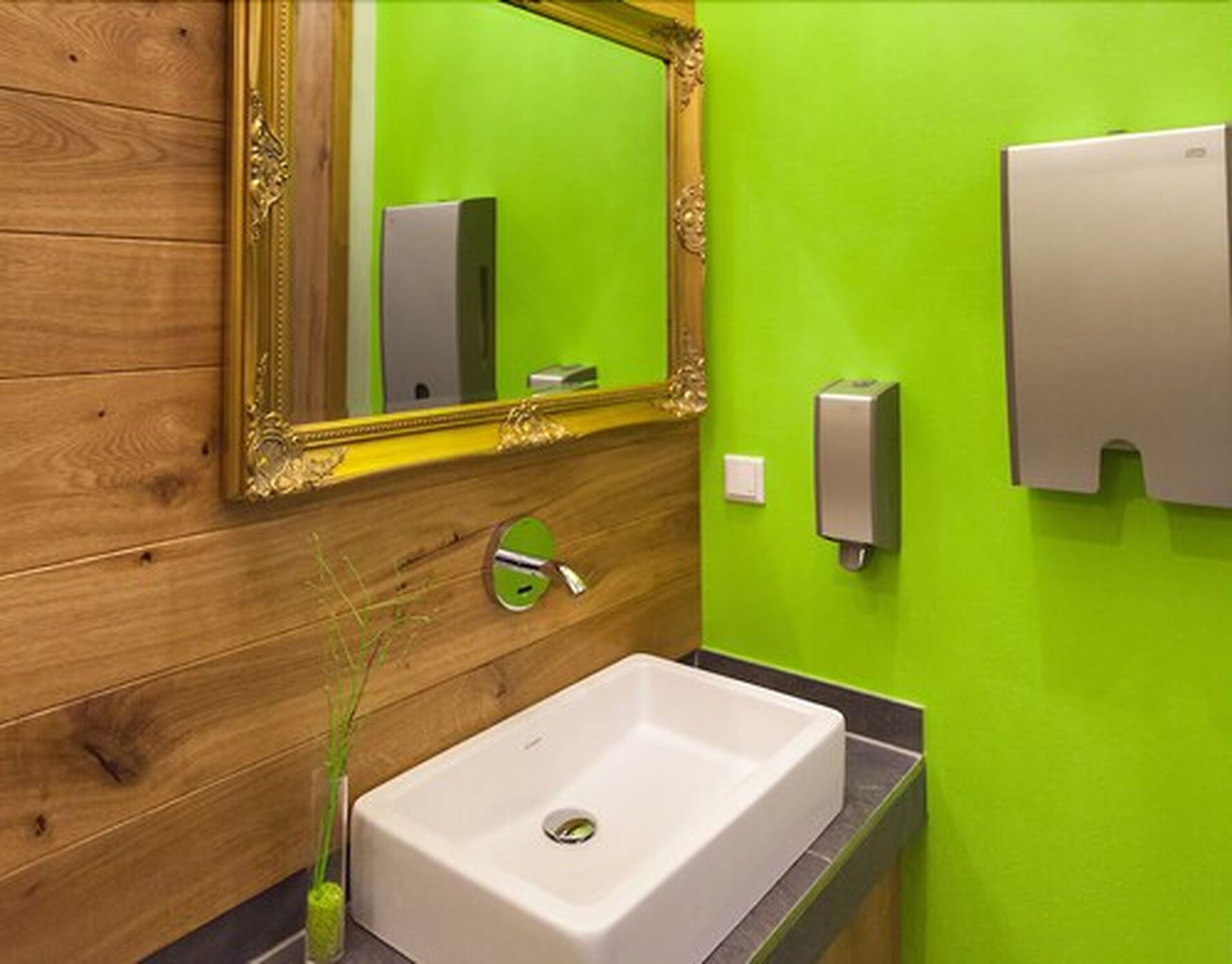 A bathroom with a wooden vanity, a white sink, a green wall, a gold-framed mirror, and a soap dispenser.