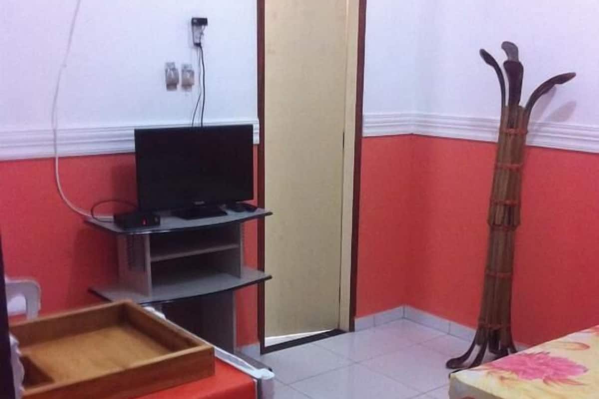 Ein kleines Zimmer mit einem Bett, einem Fernseher auf einem Schrank, einem Holzstuhl und einem Spiegel.