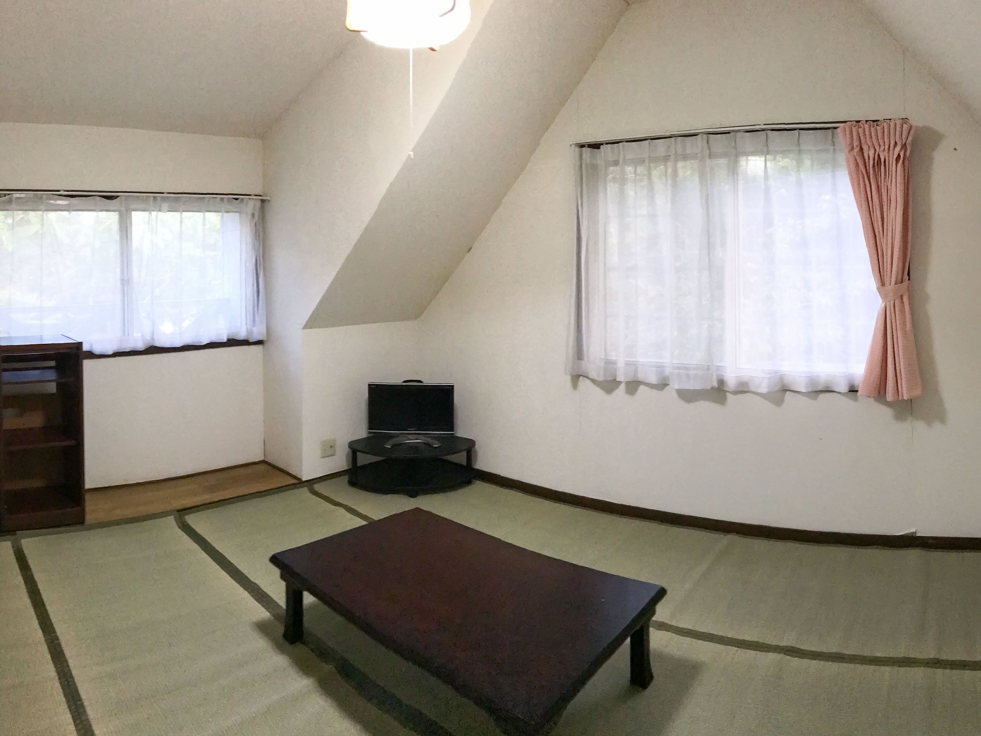 A room with a sloped ceiling, a bench, a TV stand, and a window with a curtain.