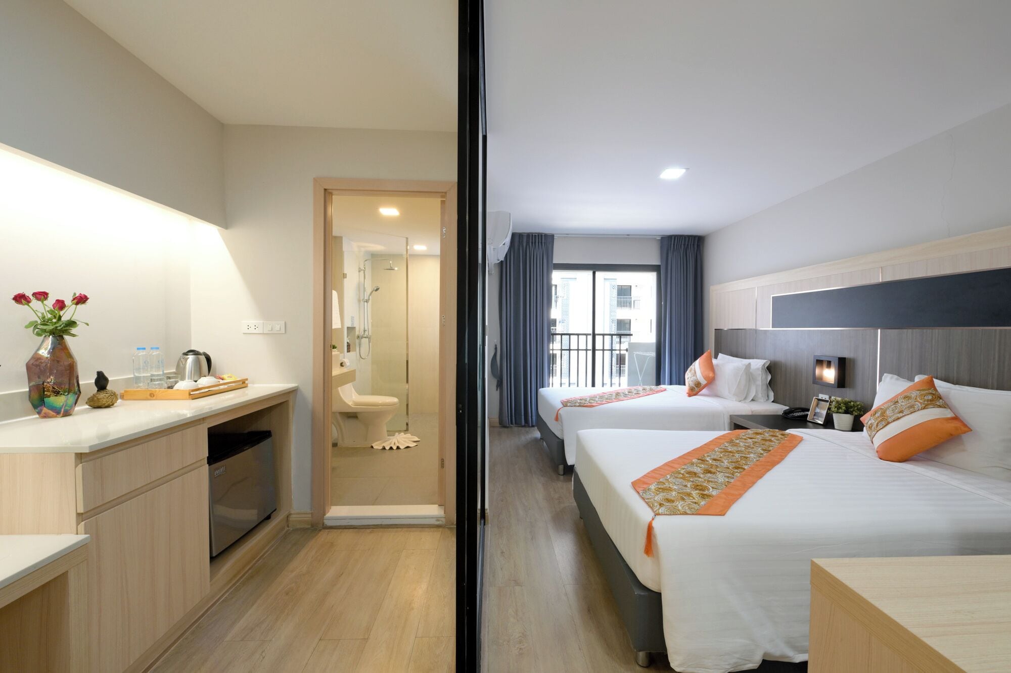 A hotel room with a kitchenette, a shower area, two beds with orange accents, and a view of the outside.