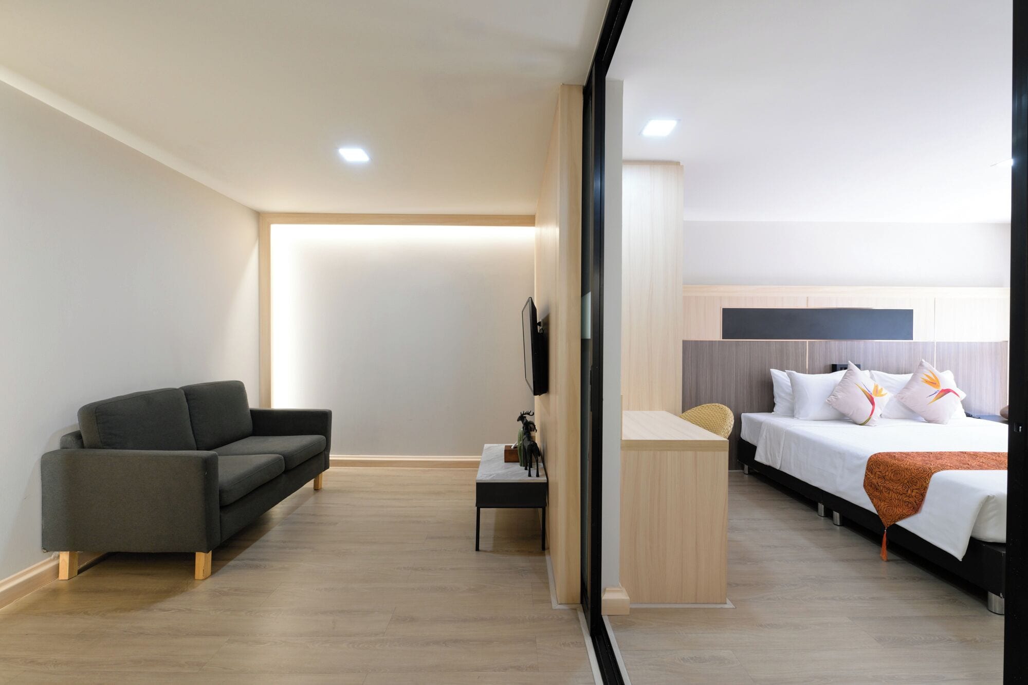 A modern hotel room with a sofa, a small table, a bed with pillows, and a TV mounted on the wall.