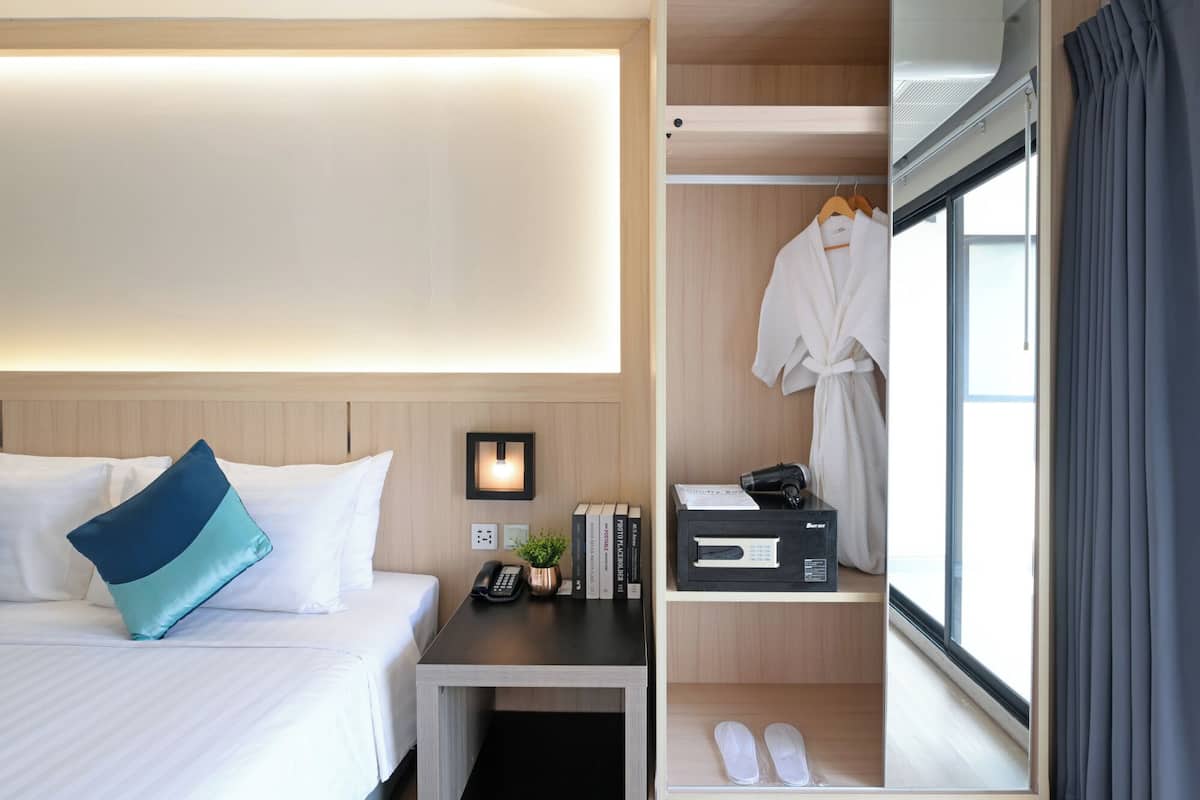 A hotel room with a bed, bedside table, lamp, books, a robe, and a shoe rack.