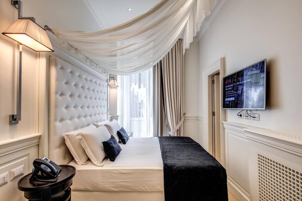 A hotel room with a tufted headboard, a bed with white and black bedding, a bedside telephone, a television on the wall, and sheer drapes.